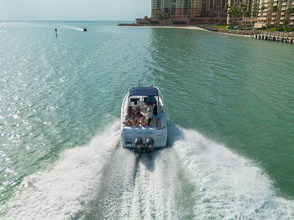 The Sweet Melissa seen from above and behind, throwing a wide wake across green water, with the Cape Marco towers on the Marco Island shoreline ahead of her